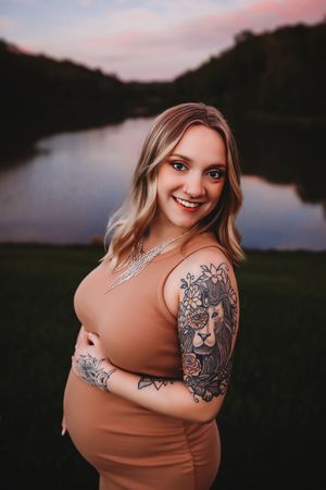 Maternity photo series at sunset showing a woman in brown dress with shoulder tattoos by a lake.