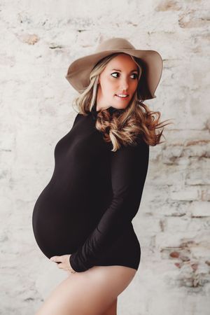 A maternity photo series showing a woman in black dress and beige hat posing against a white brick wall.