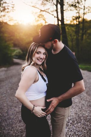 A couple shares an intimate moment on a gravel path at sunset with golden light filtering through trees in the background.