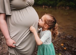 A maternity photo shoot by a creek shows a tender embrace between a pregnant woman and a child in green dress.