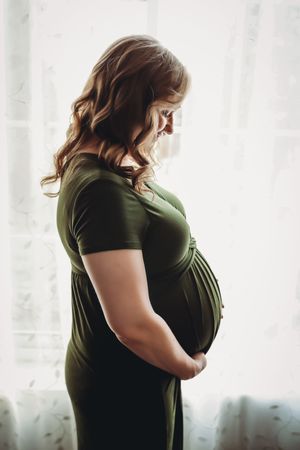 Side view silhouette of a pregnant woman in an olive green shirt standing against a bright window background.