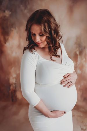 Maternity portrait of an expectant mother in a white fitted dress against a brown backdrop.