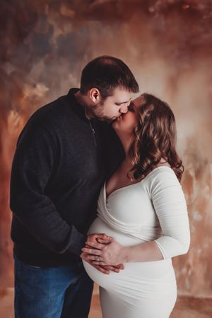 Intimate maternity photo of couple sharing a tender kiss against a warm brown backdrop.