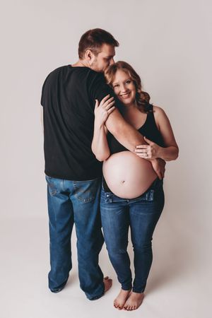 Intimate maternity photo of expecting couple embracing and smiling against neutral background.