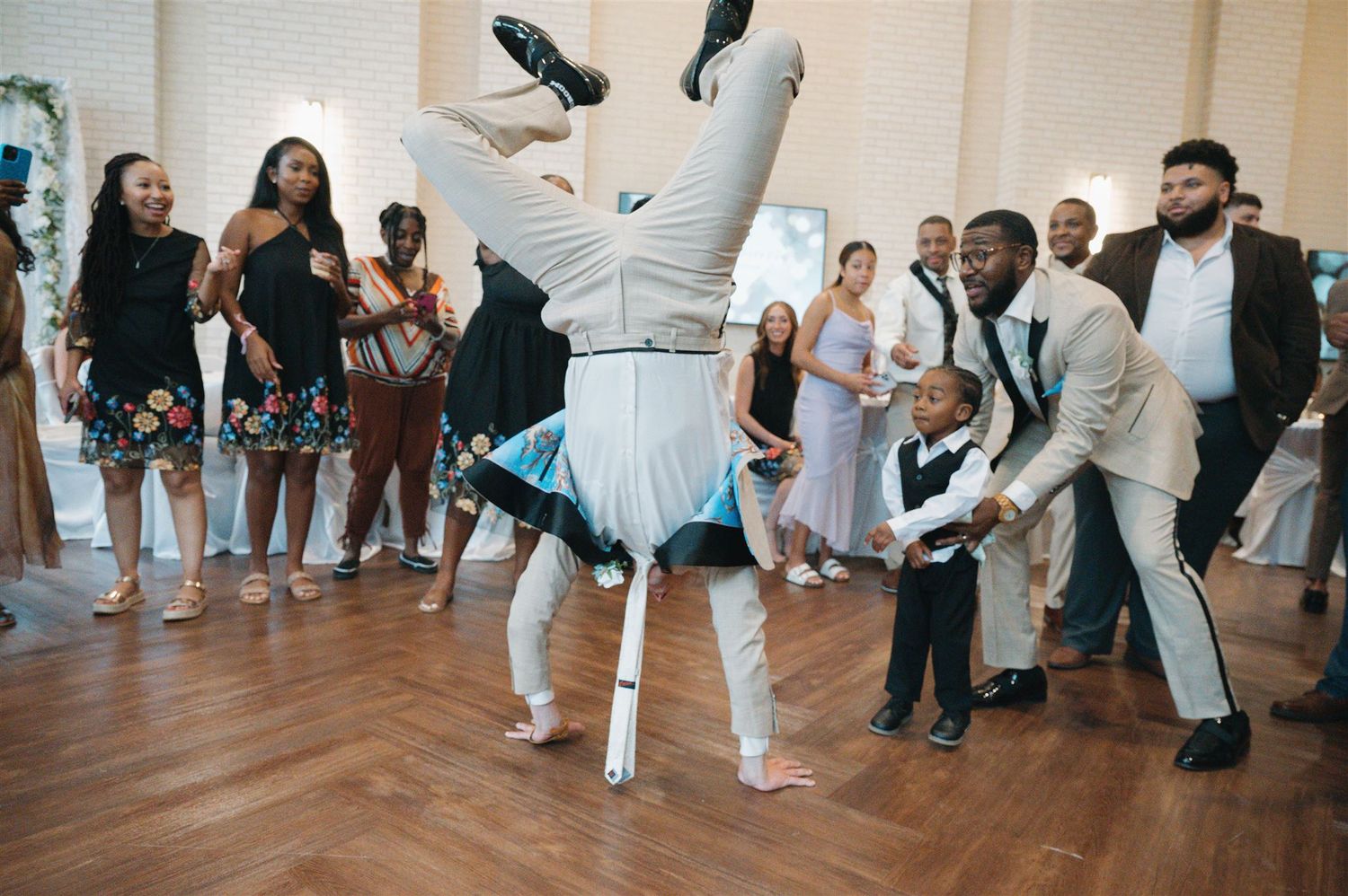 Dancer performs dramatic headstand in blue tutu at party while onlookers react with excitement on hardwood floor.