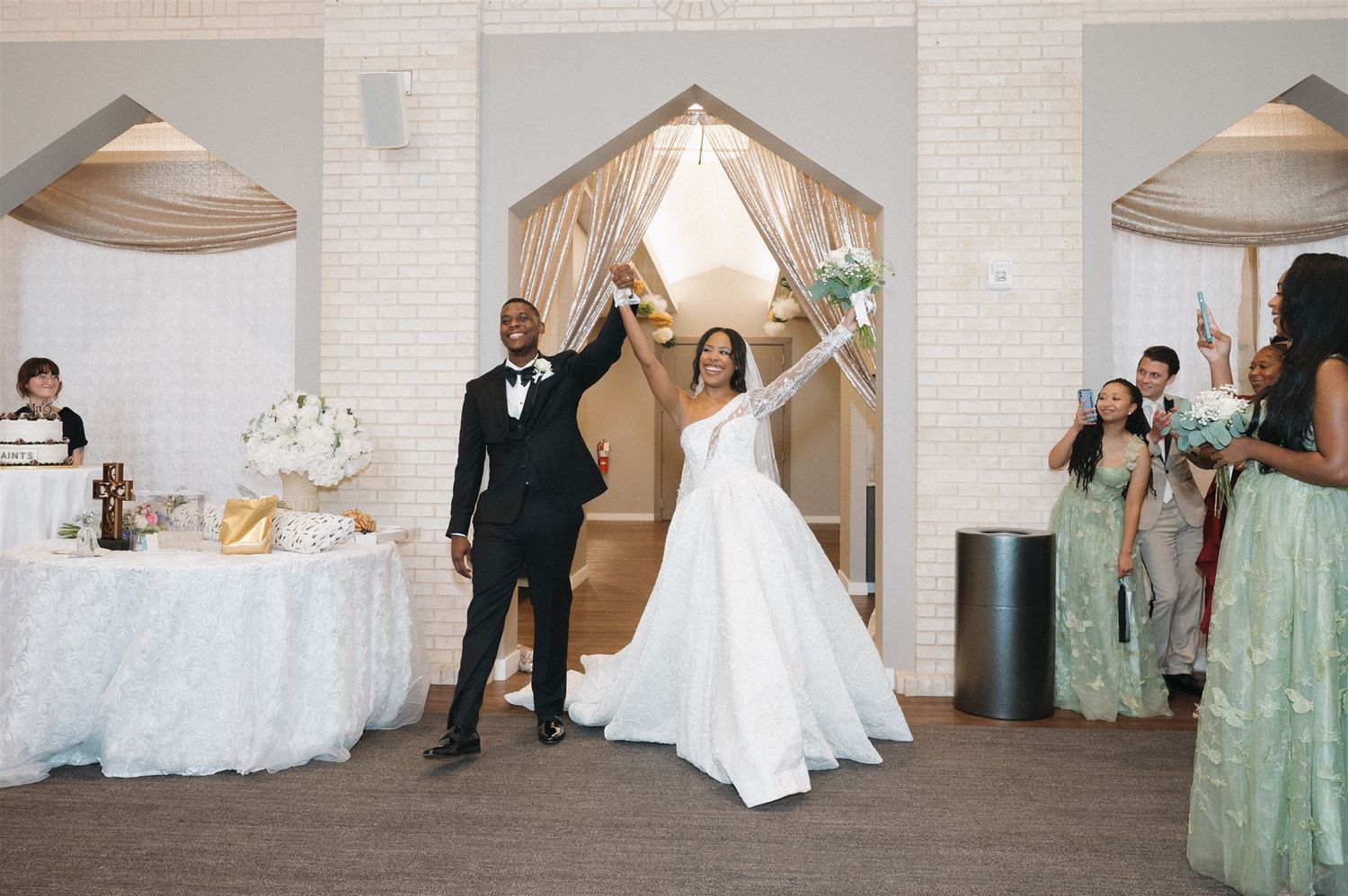 A joyful newlywed couple celebrates during their wedding reception in an elegant white-walled venue with decorative archways.