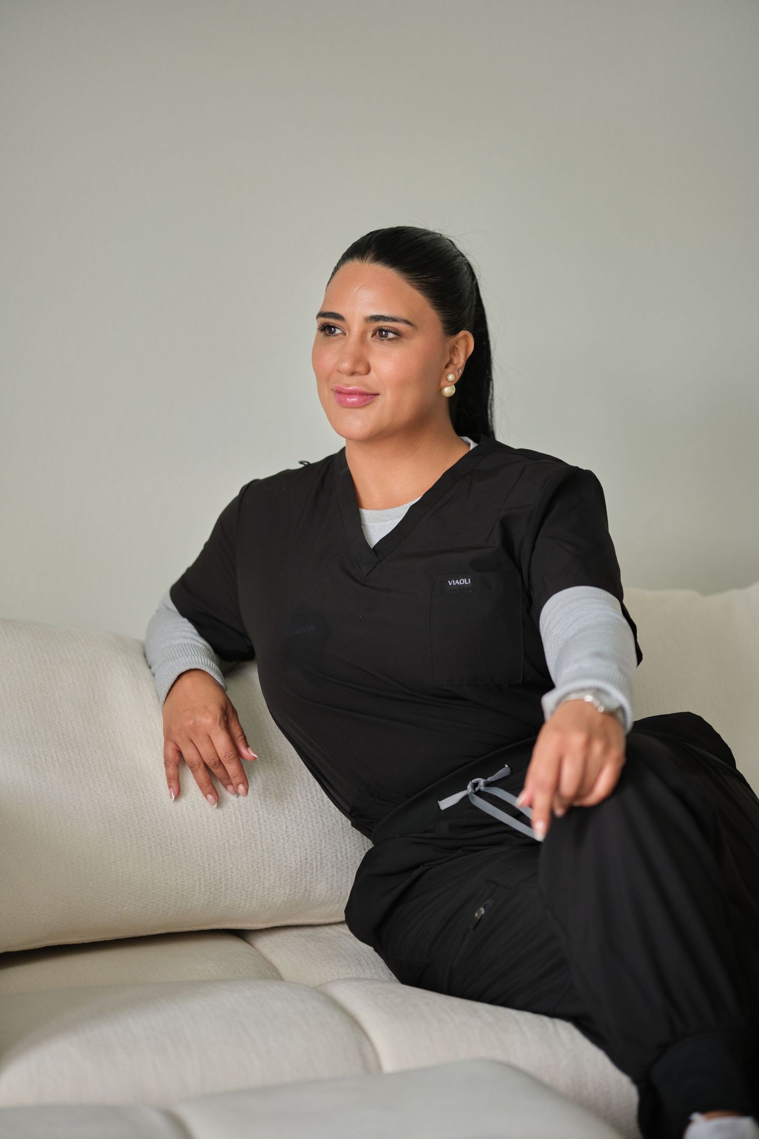 A healthcare professional in black scrubs sits relaxed on a white sofa in a modern clinical setting.