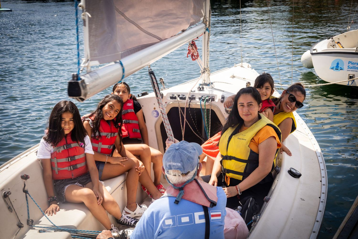 A group enjoying a sunny day on a sailboat wearing red and yellow life jackets on calm waters.