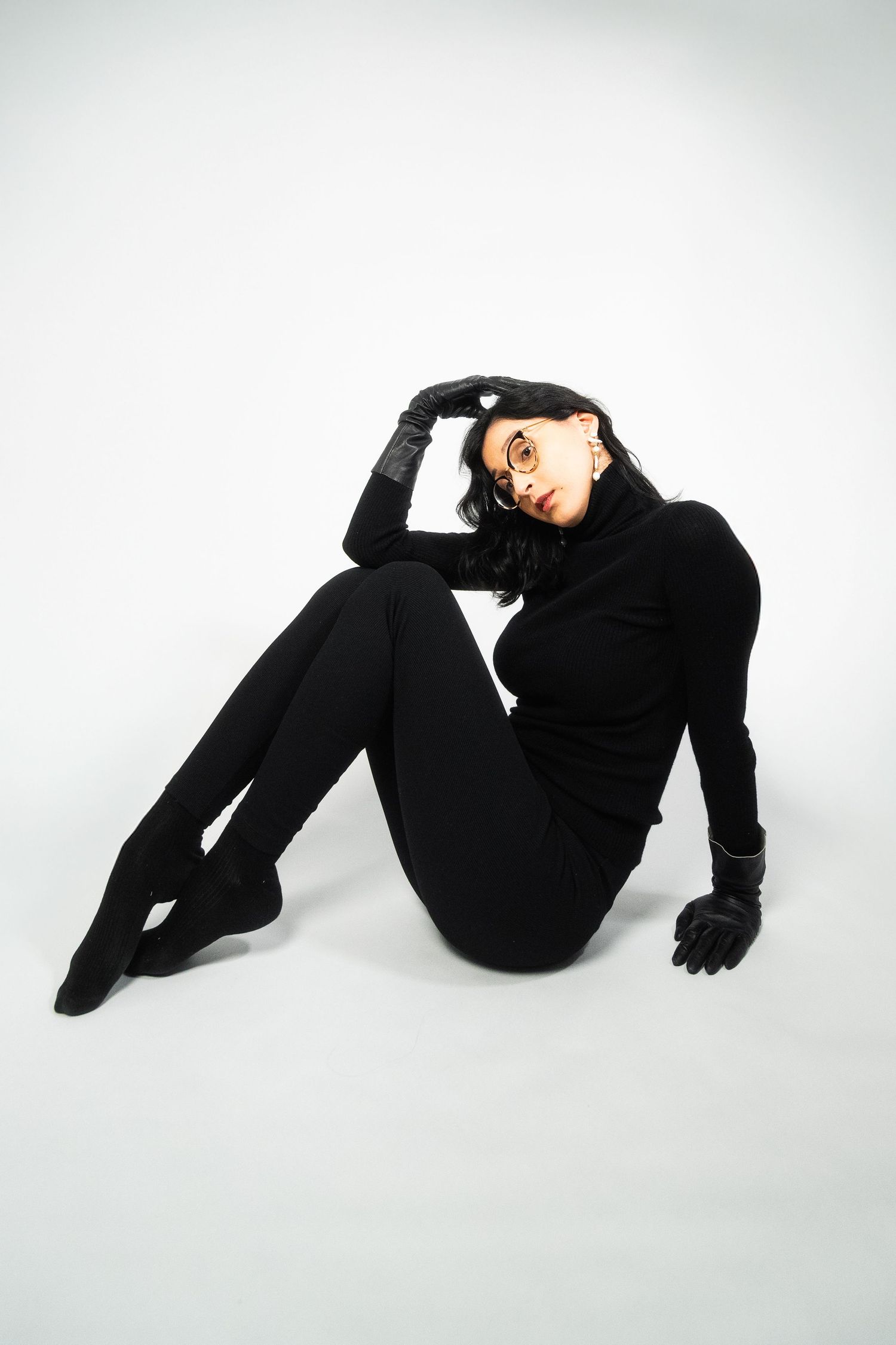 Person in all black outfit lying on white background in artistic pose.