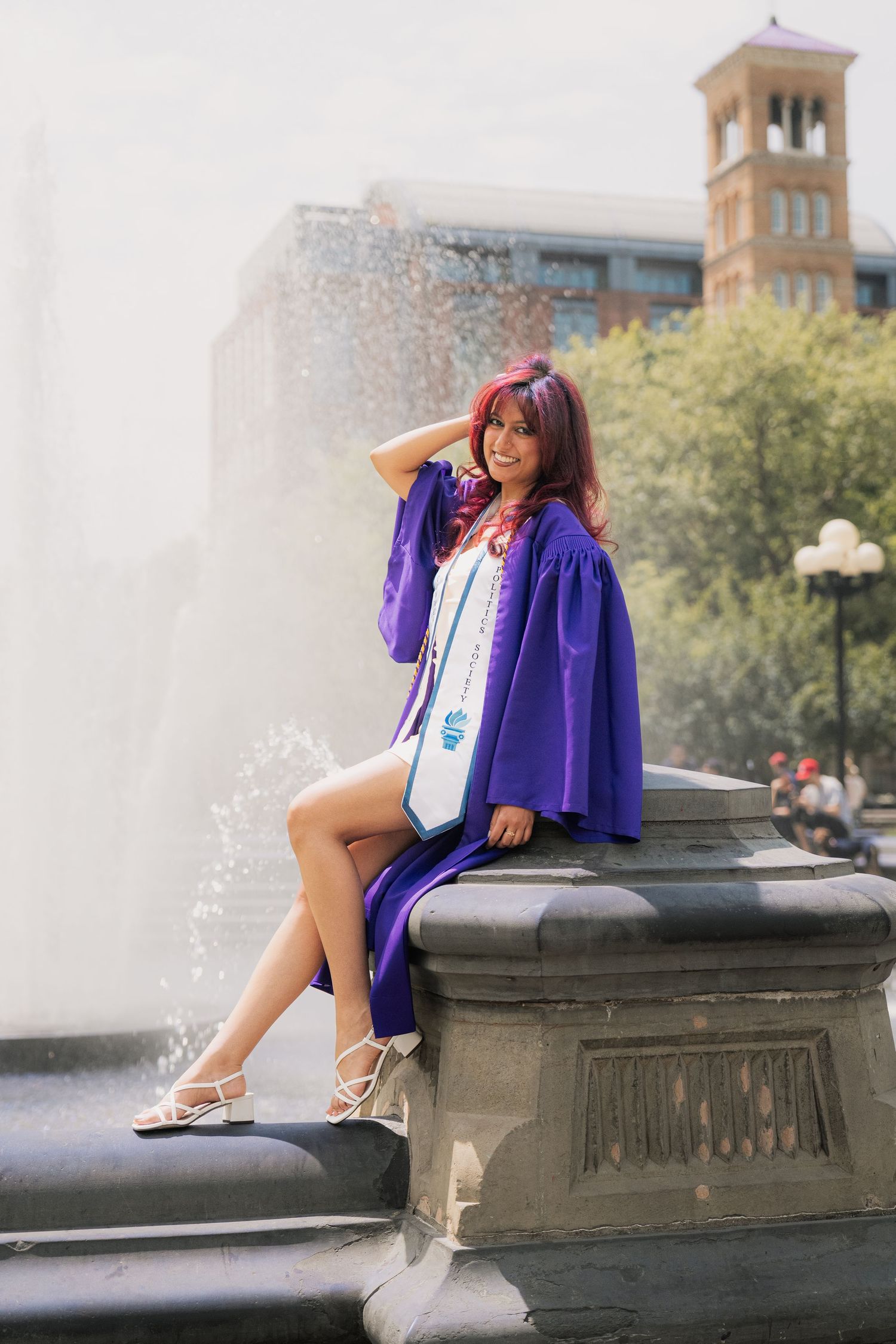 Person in purple graduation gown posing playfully by fountain on campus grounds.