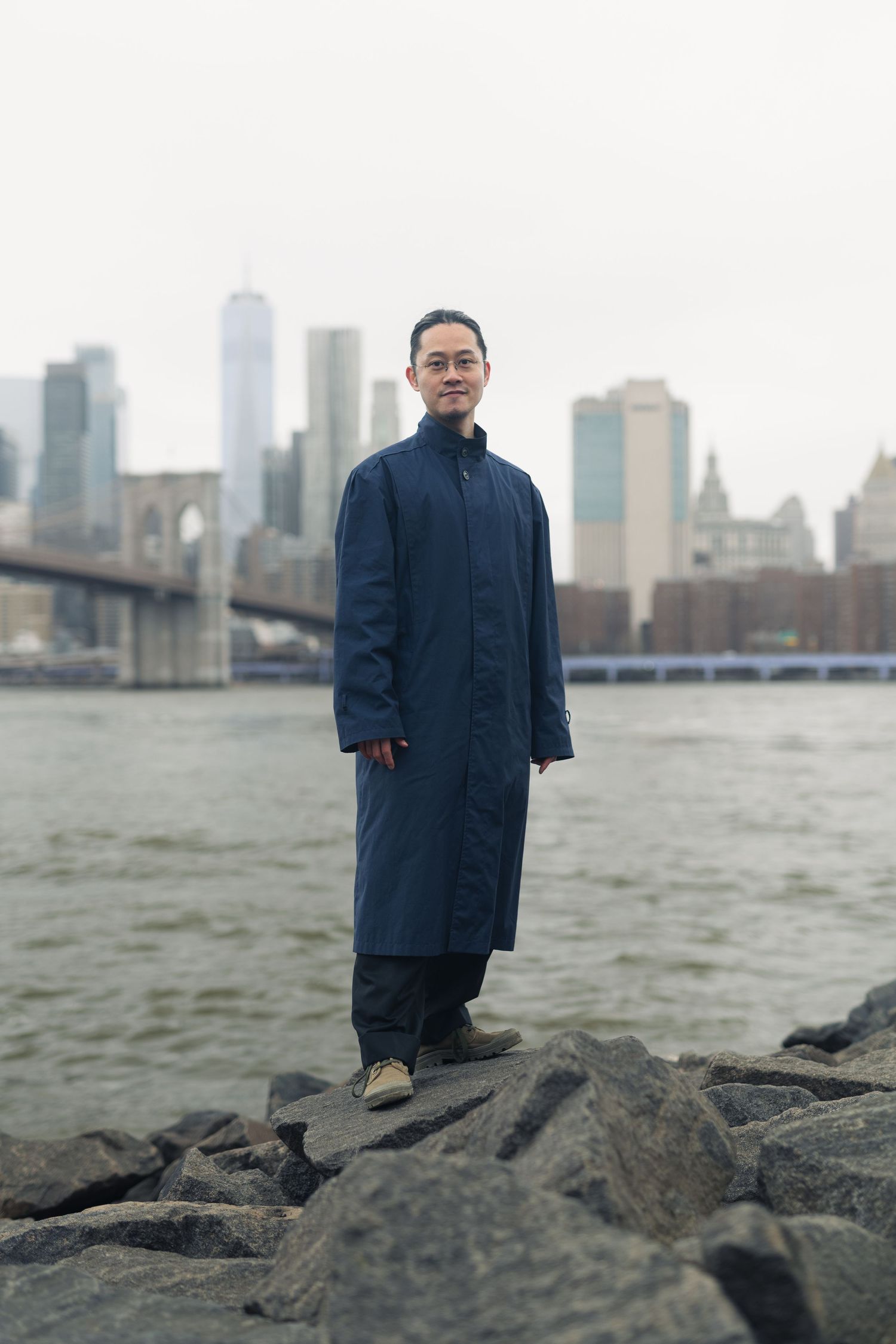 A figure in a dark coat stands on rocky shoreline with city skyline in background.
