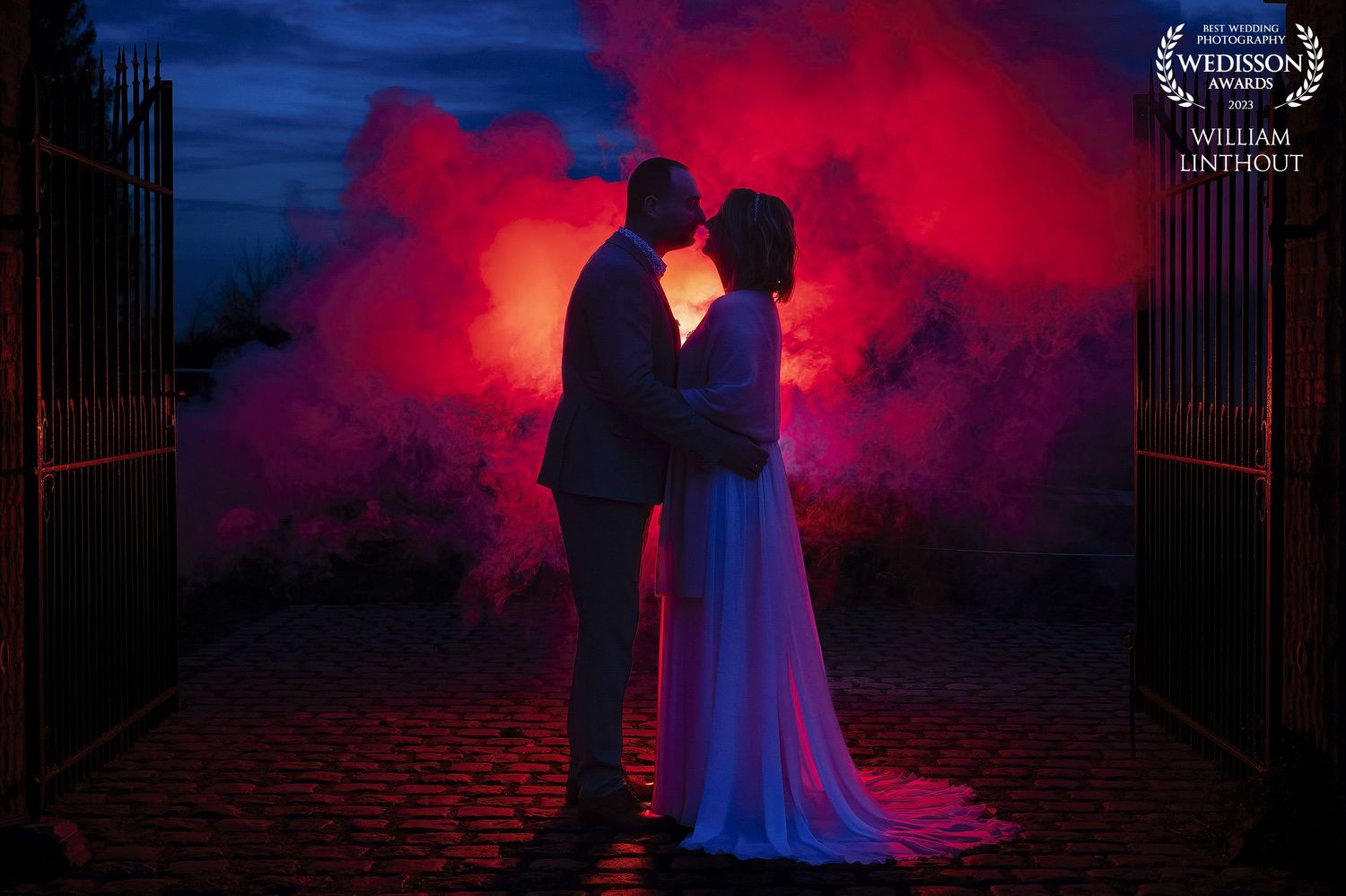 Silhouetted couple against dramatic red smoke and blue sky backdrop in romantic wedding photo.