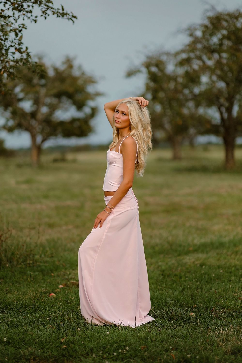 Blonde woman in pink crop top and maxi skirt posing outdoors in a green field with trees in background.
