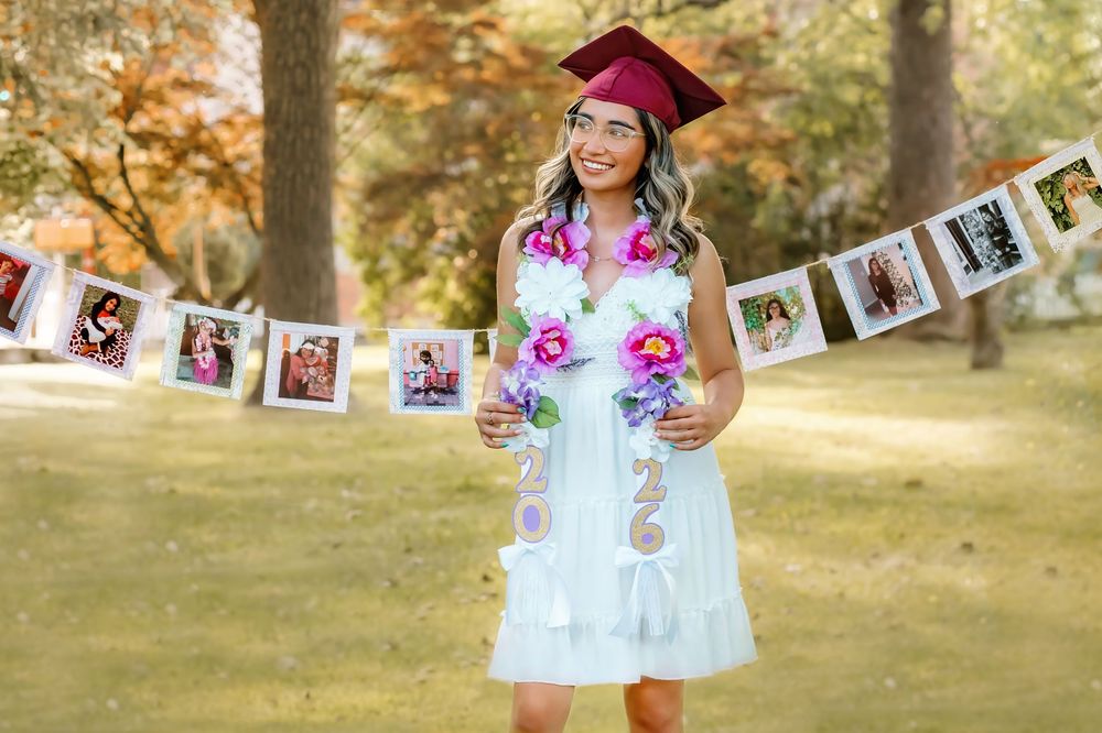 Happy graduate in white dress and maroon cap posing with 2026 decorations and photo banner in park setting.