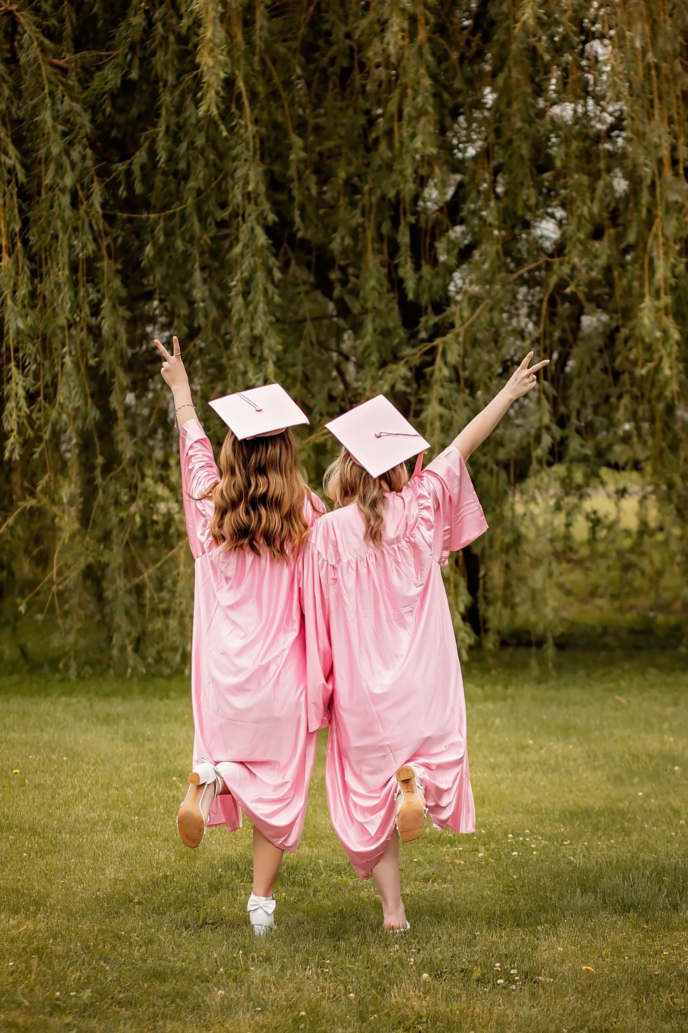 Two female graduates in pink cap and gown jumping with arms raised, viewed from behind outdoors.