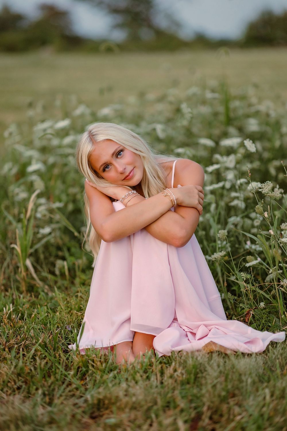 Blonde girl in pink dress sitting in wildflower field hugging her knees.