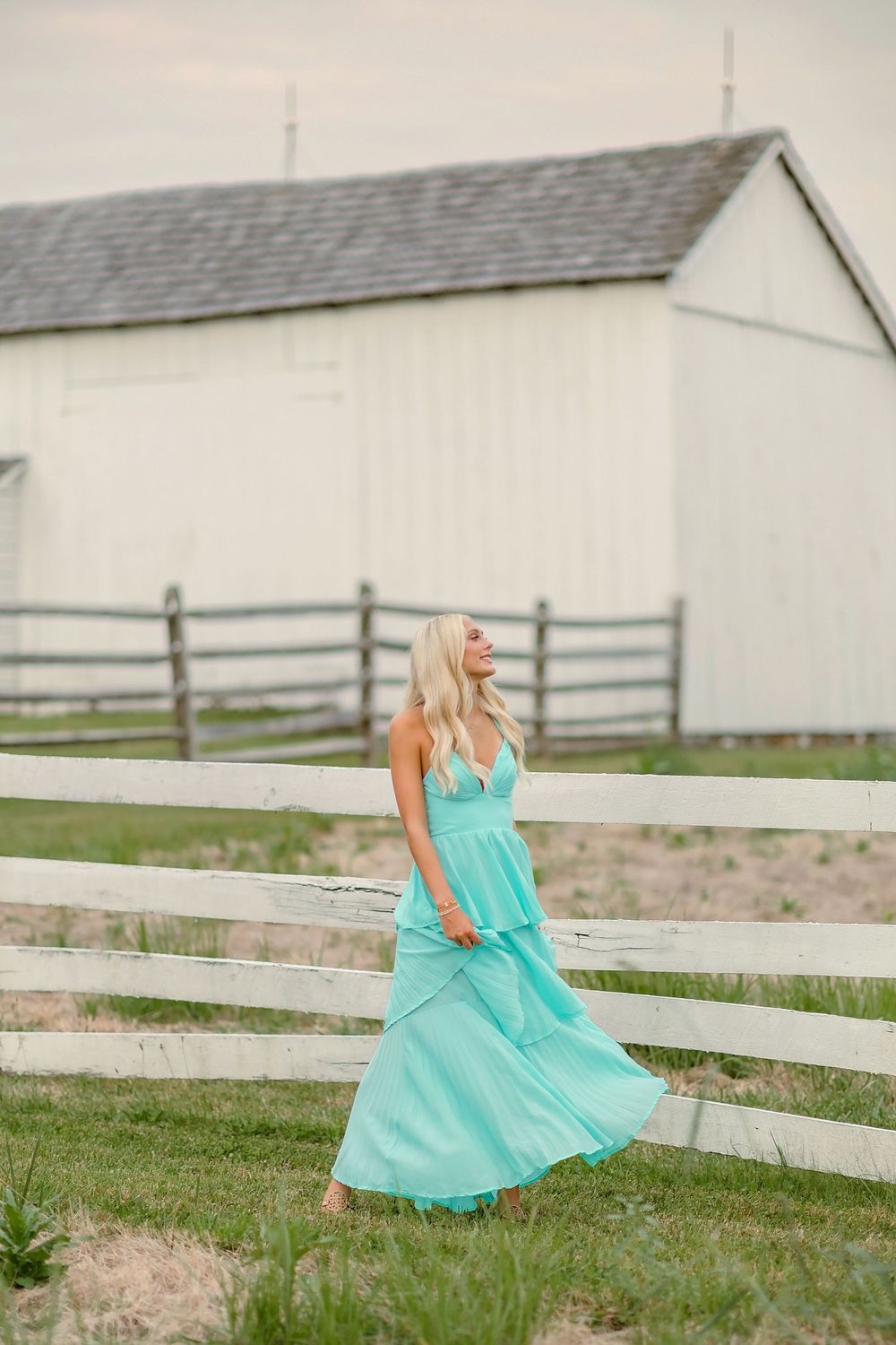 Blonde woman in flowing teal maxi dress walking by white wooden fence on a farm.