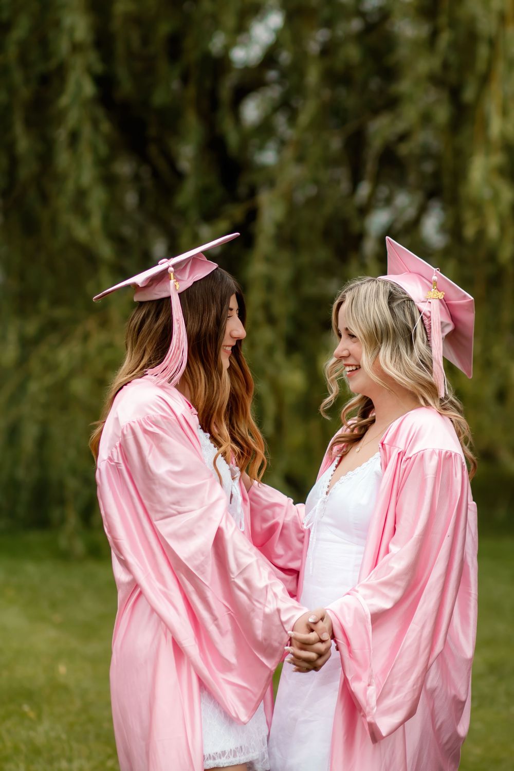 Two female graduates in matching pink cap and gown holding hands and smiling at each other outdoors.