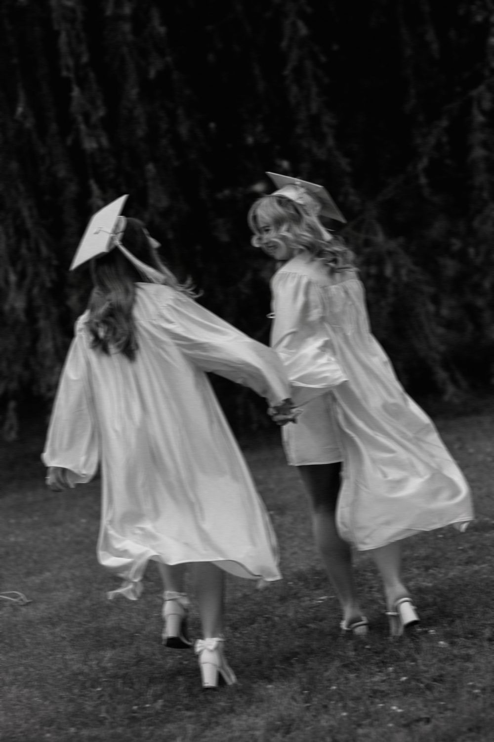 Black and white photo of two female graduates in white cap and gown holding hands walking outdoors.