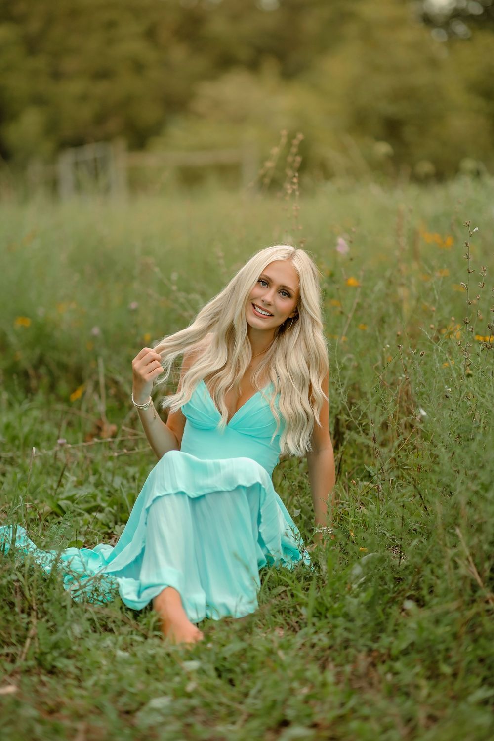 Smiling blonde woman in a flowing teal dress sitting gracefully in a lush green meadow with wildflowers.
