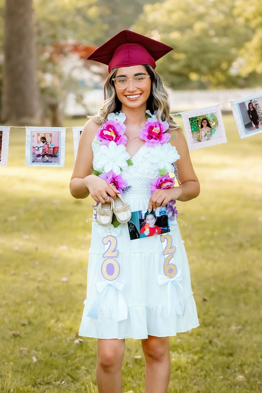 Smiling graduate in white dress and maroon cap wearing floral lei with 2026 decorations at outdoor graduation photo session.