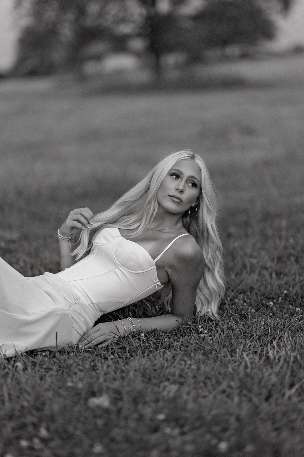Black and white photo of a blonde woman in a white dress lying gracefully on green grass in an outdoor field.