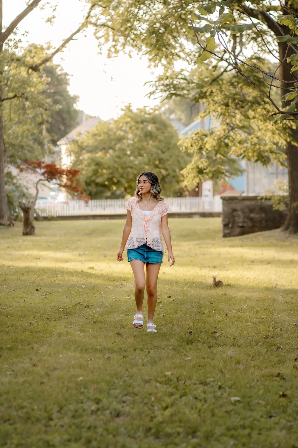 Young woman in pink top and denim shorts walking through lush green park on a sunny summer day.