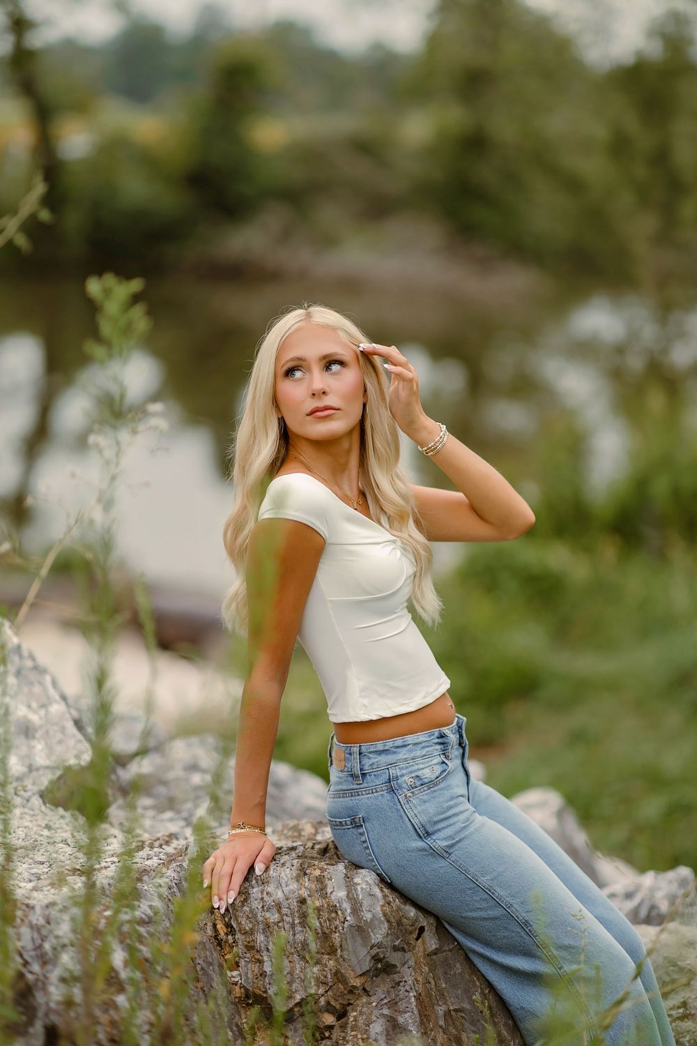 Blonde woman in white crop top and jeans poses on rocks near a river in a natural outdoor setting.