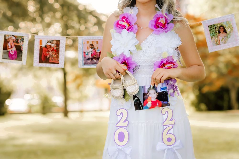 Close-up of graduate in white dress displaying 2026 decorations, floral lei, and holding photos at outdoor celebration.