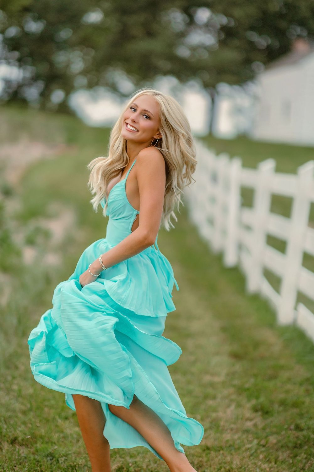 Smiling blonde woman twirling in a flowing turquoise dress beside a white fence on a green farm.