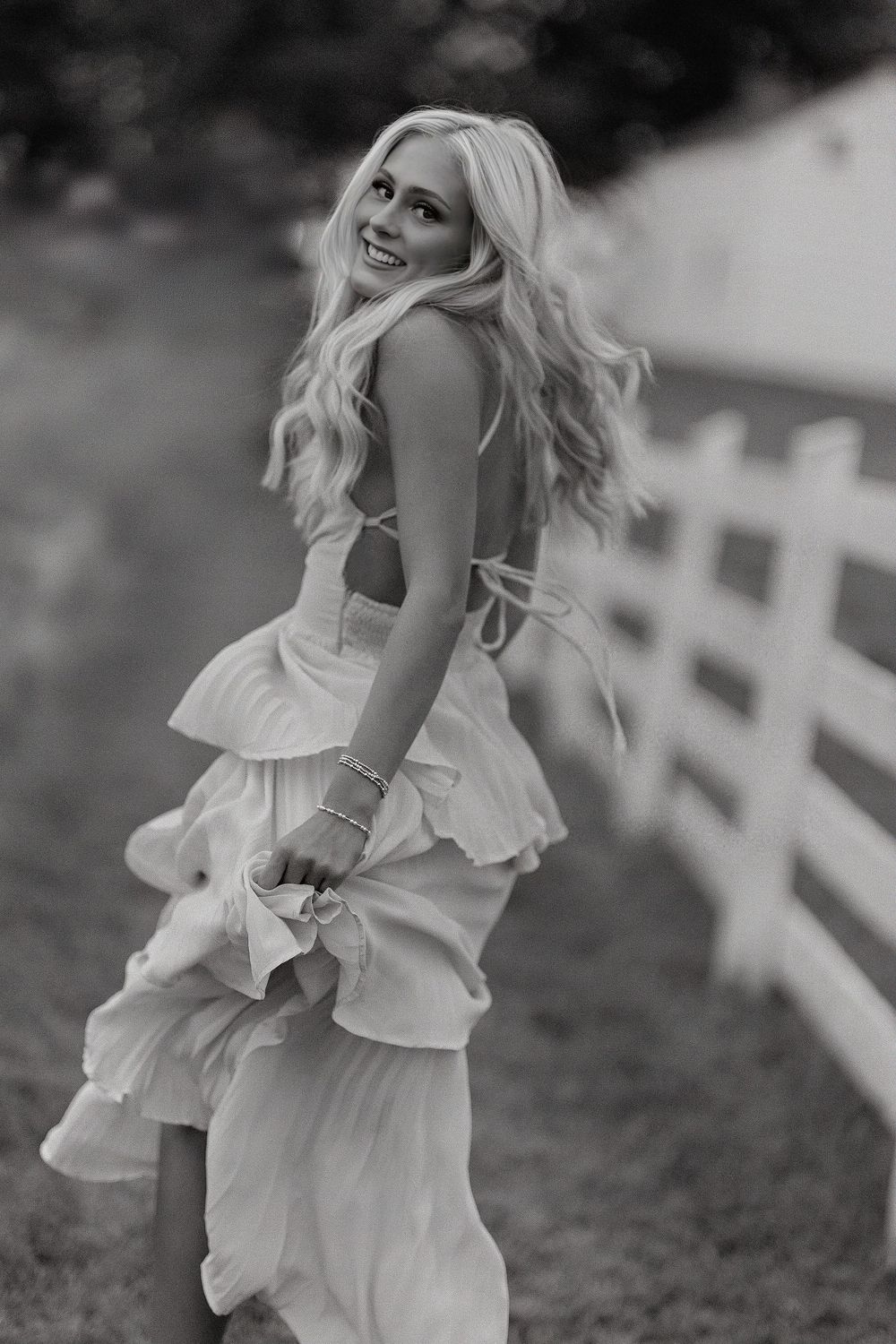 Smiling blonde woman in a ruffled backless white dress twirling near a white fence in a black and white photo.