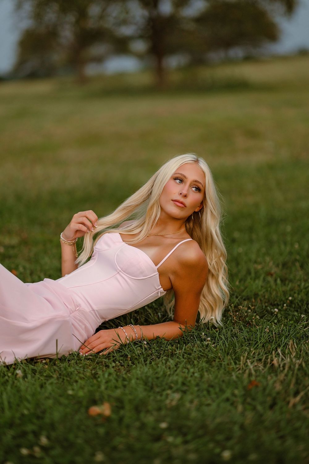 Young blonde woman in pink dress lying gracefully on green grass, gazing upward in a serene outdoor setting.