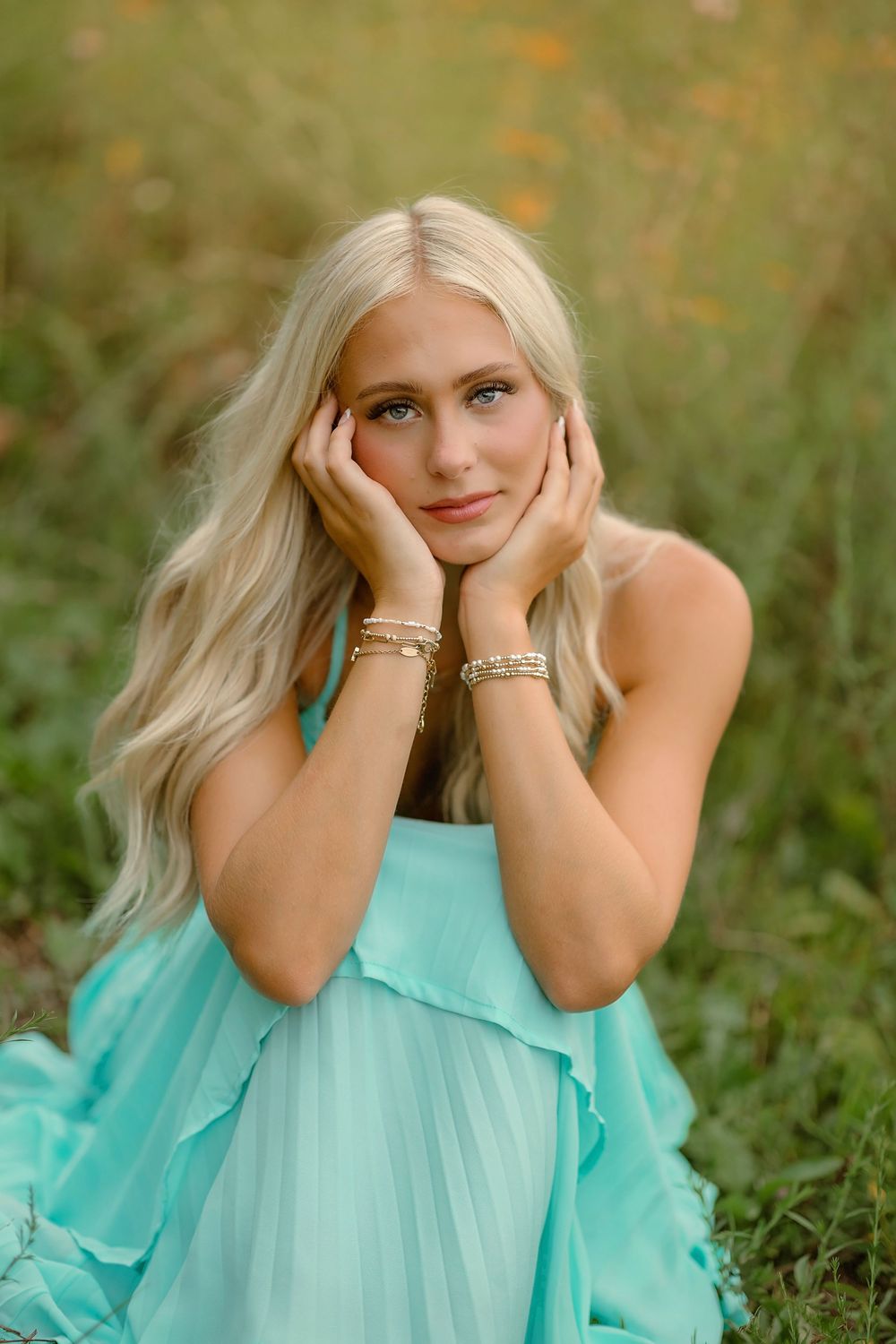 Blonde woman in mint dress sitting in green field, hands framing face, wearing gold bracelets, soft natural light.