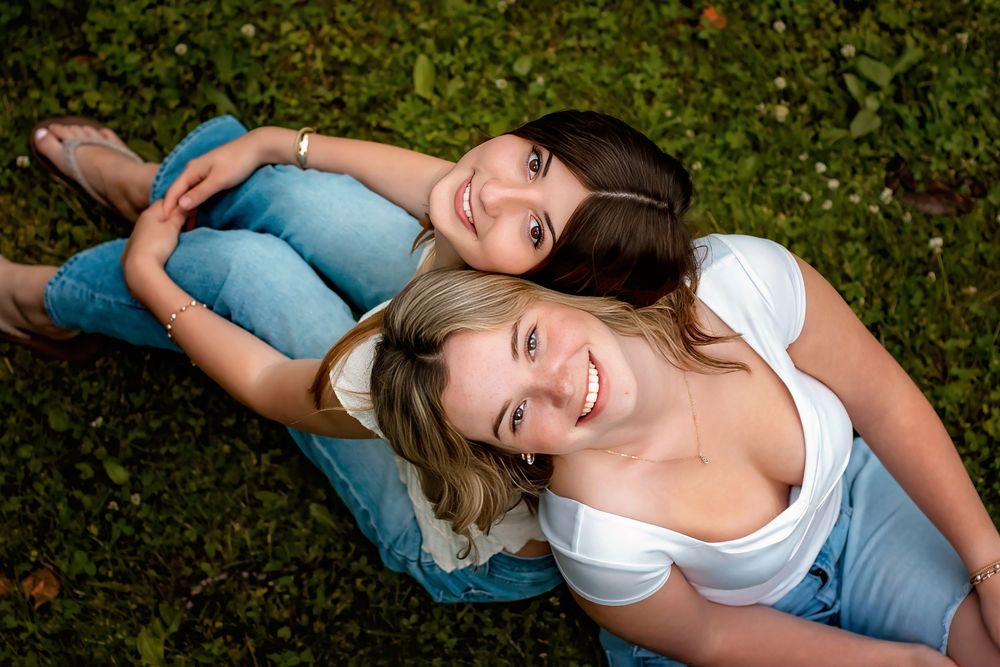 Two smiling young women lying on green grass, looking up at camera in a relaxed outdoor portrait.
