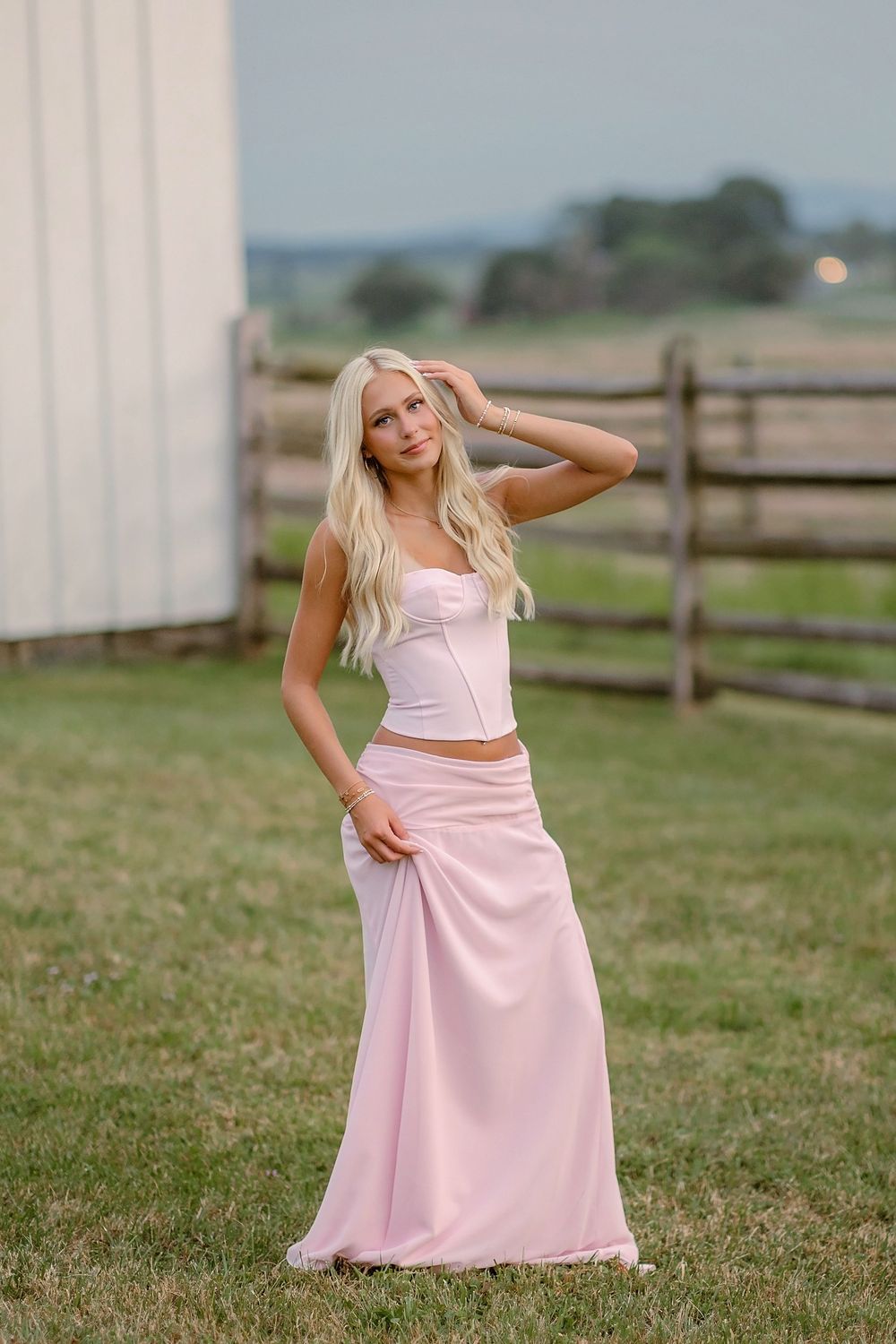 Blonde woman in white corset top and pink maxi skirt poses gracefully near a rustic fence on a farm.