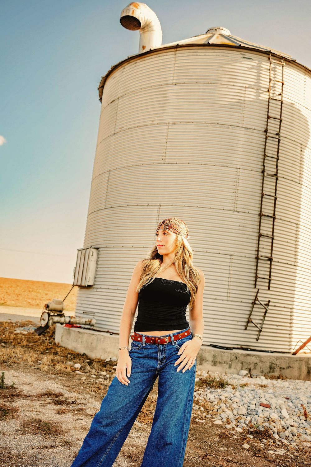 Young blonde woman in black top and wide-leg jeans poses confidently in front of a large grain silo on a farm.