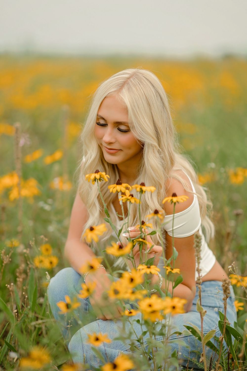 Blonde woman crouching in a vibrant yellow wildflower field, gently touching black-eyed Susan flowers.