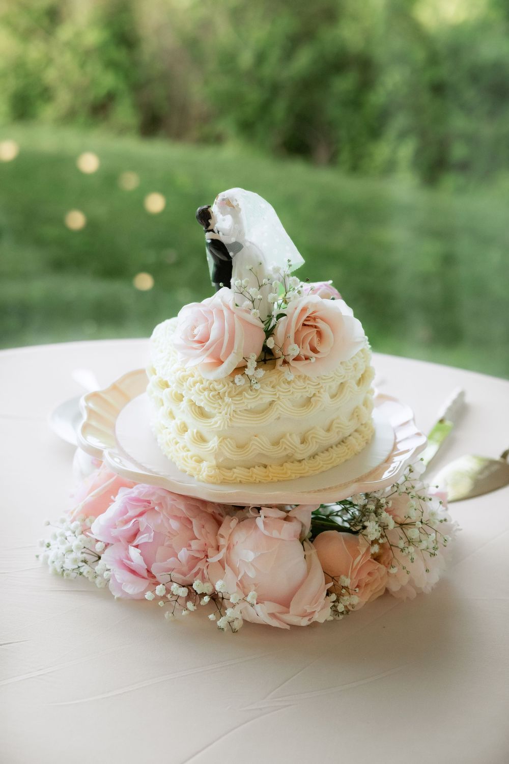 Elegant one-tier white wedding cake with pink roses, bride and groom topper, surrounded by peonies and baby's breath. Wedding Photography