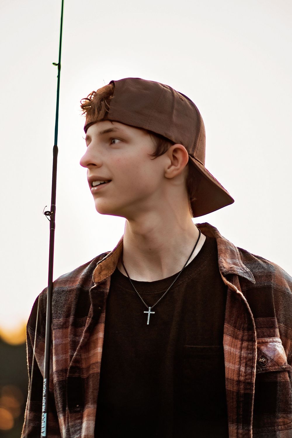 Teenage boy wearing backward cap and cross necklace holding a fishing rod at sunset. Guy Codorus Fall Senior Pictures