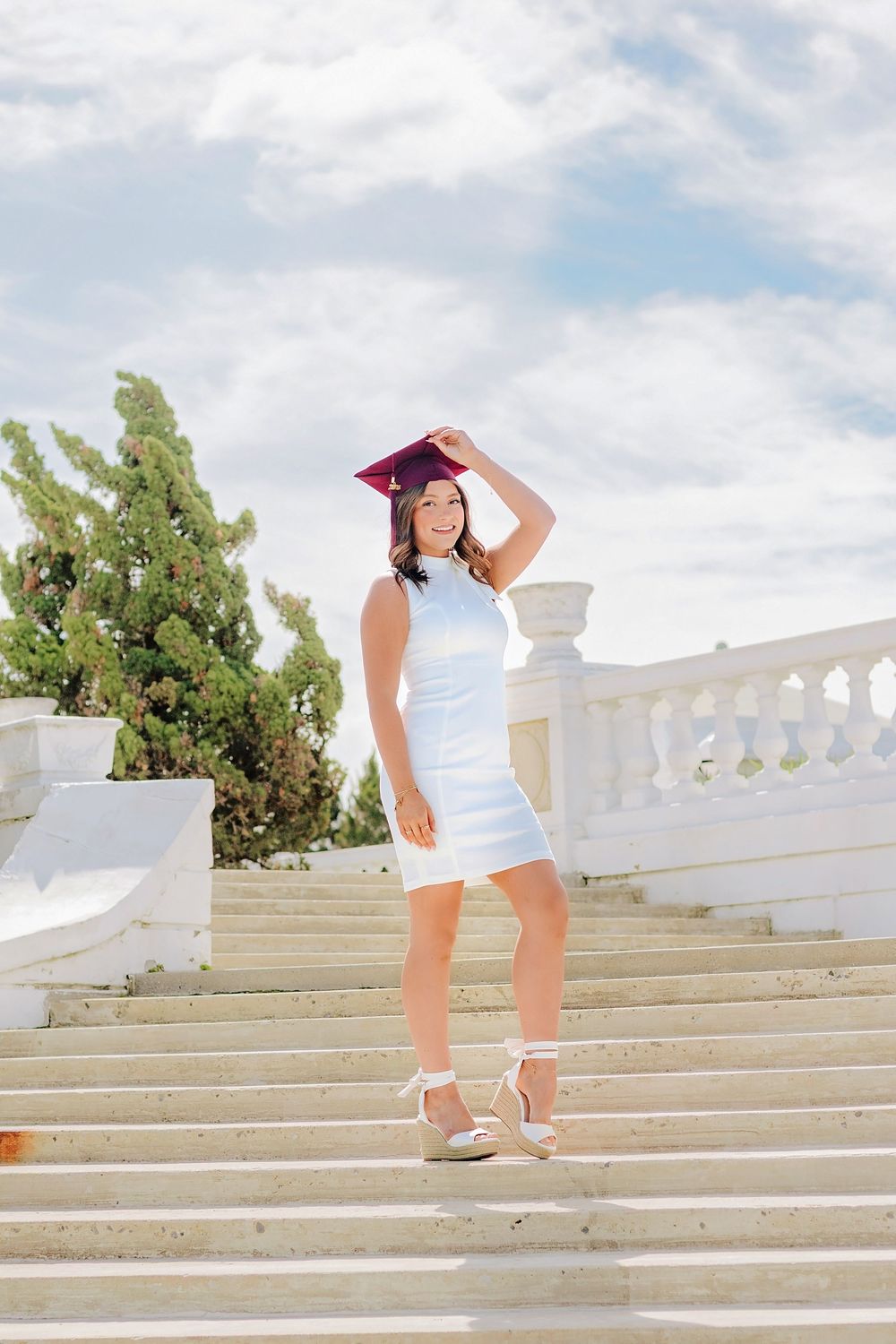 Smiling graduate in white dress and maroon cap posing on grand white marble staircase outdoors. Hershey Hotel Senior Pictures