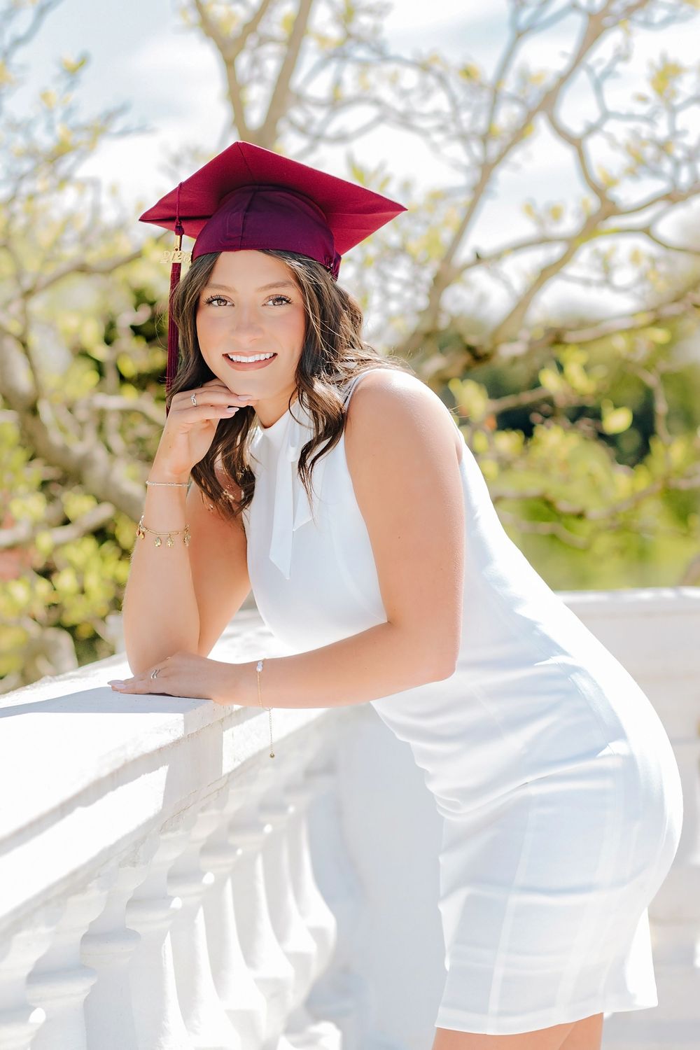 Smiling graduate in white dress and maroon cap leaning on white balcony railing outdoors. Hershey Hotel Senior Pictures