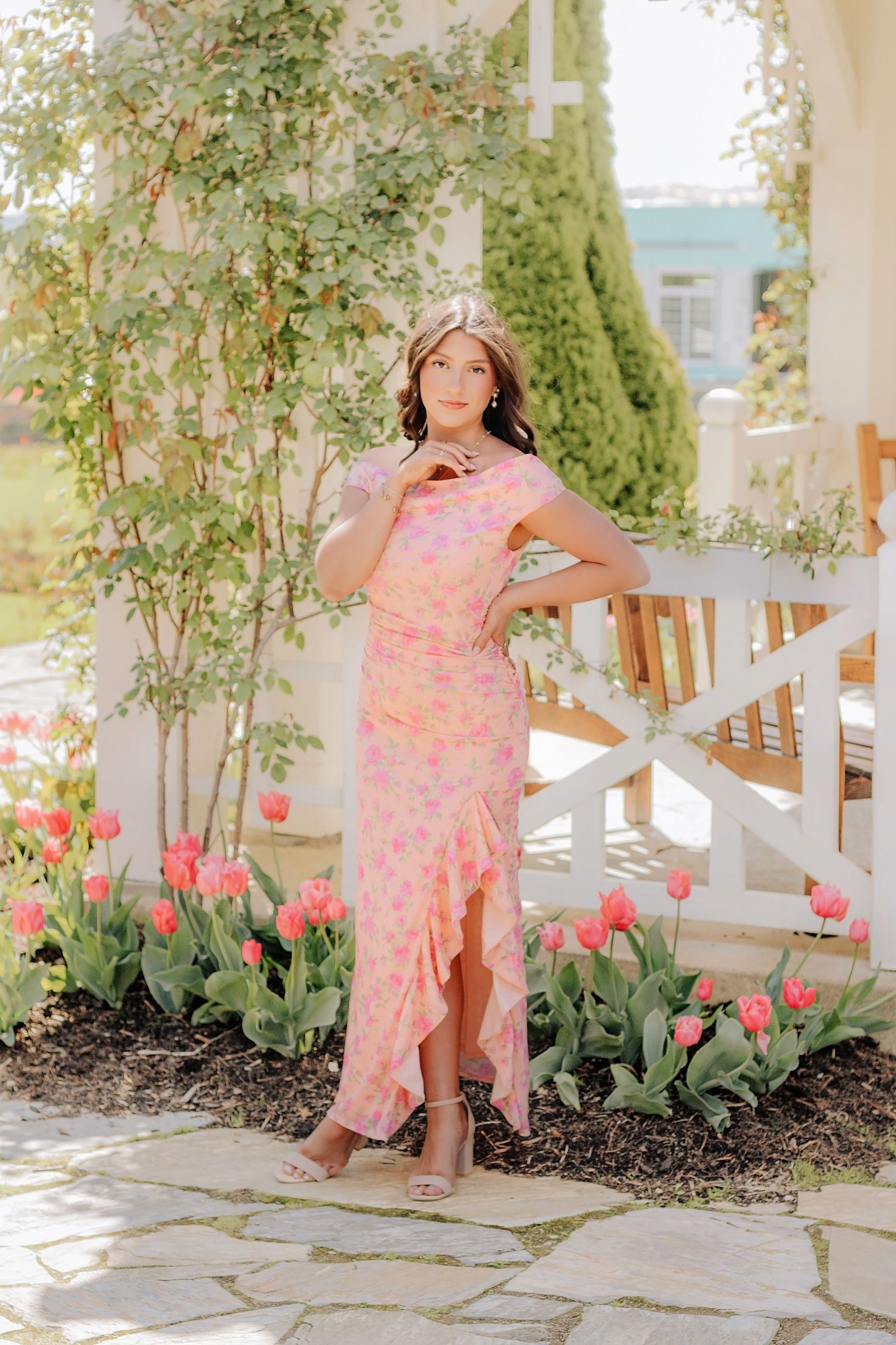 Young woman in pink floral midi dress poses among tulips on a stone patio with white pergola. Hershey Gardens Senior Pictures