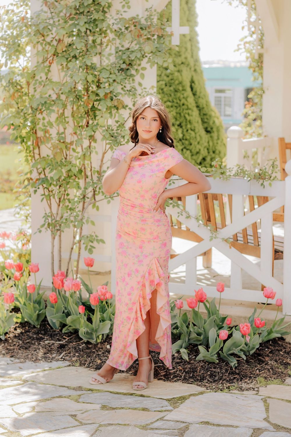 Young woman in pink floral midi dress poses among tulips on a stone patio with white pergola. Hershey Gardens Senior Pictures