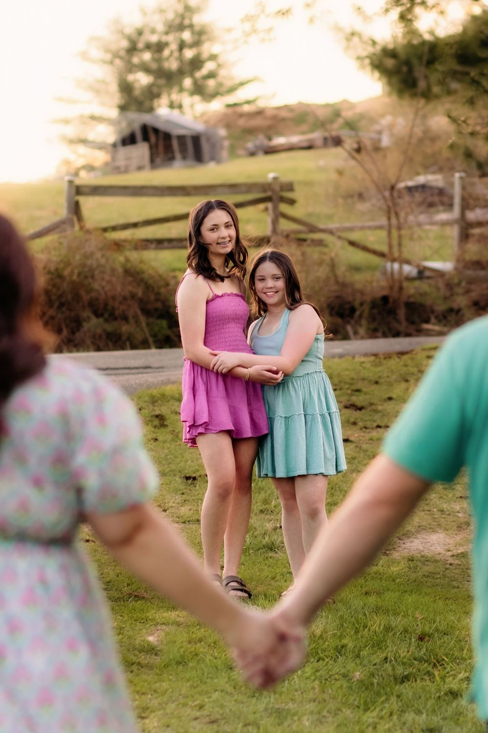 Two sisters smiling and embracing outdoors while parents hold hands in foreground during family portraits.