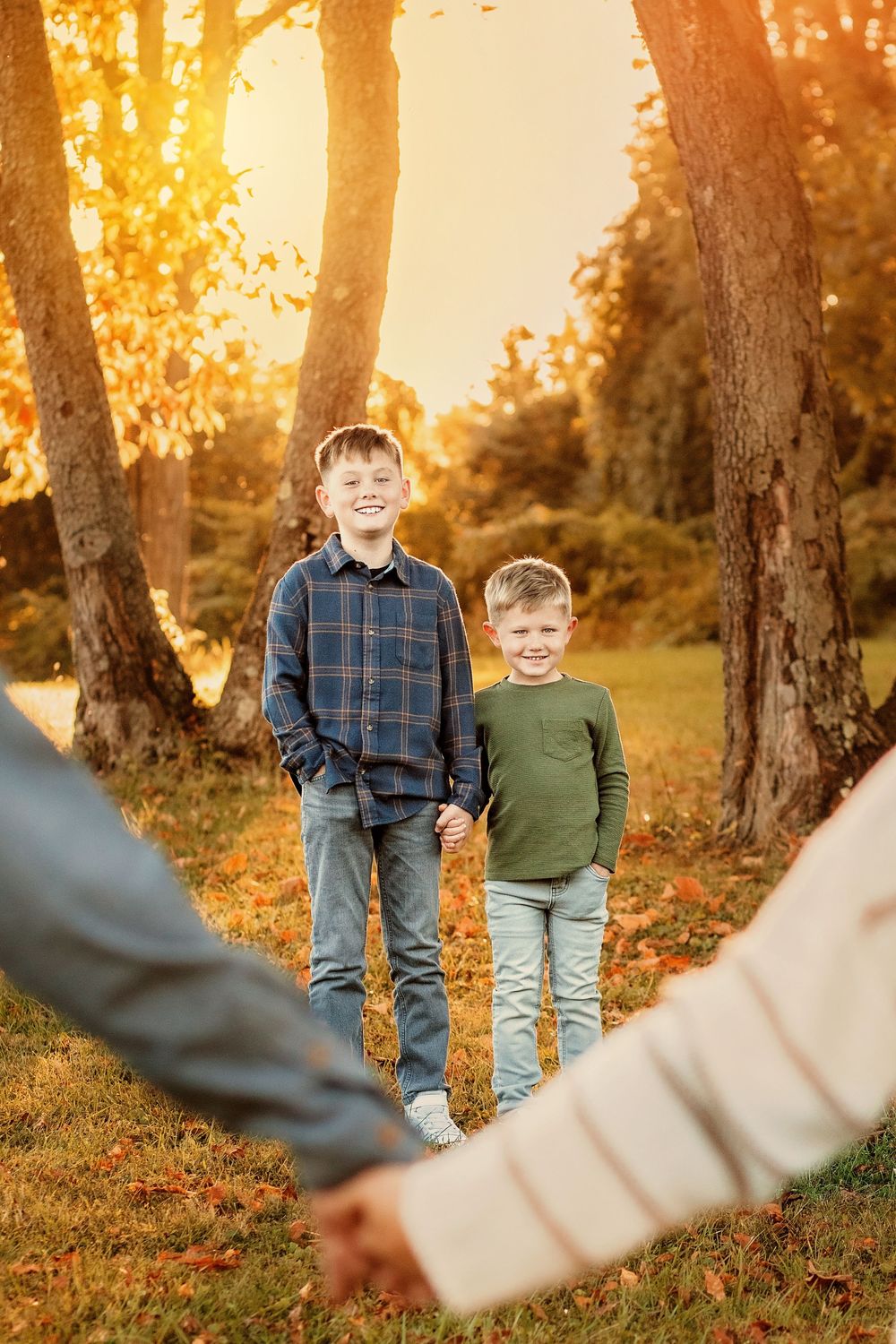 Two smiling boys hold hands in a sunlit autumn park with golden foliage. Codorus Fall Family Pictures Hanover PA