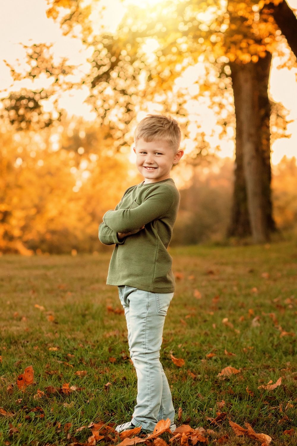 Smiling young boy with arms crossed standing in autumn park with golden foliage and fallen leaves. Codorus Fall Family Pictures Hanover PA