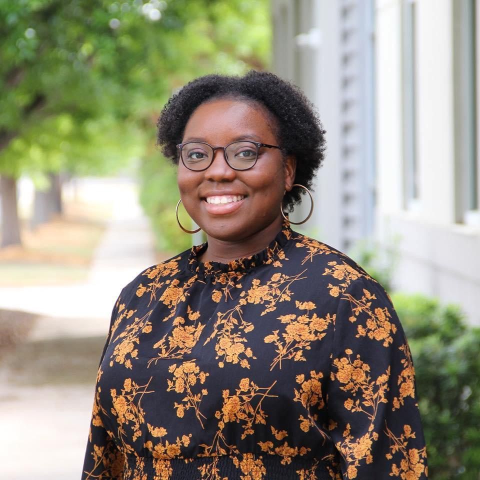 Portrait of someone wearing a floral print black and orange top with glasses and hoop earrings.