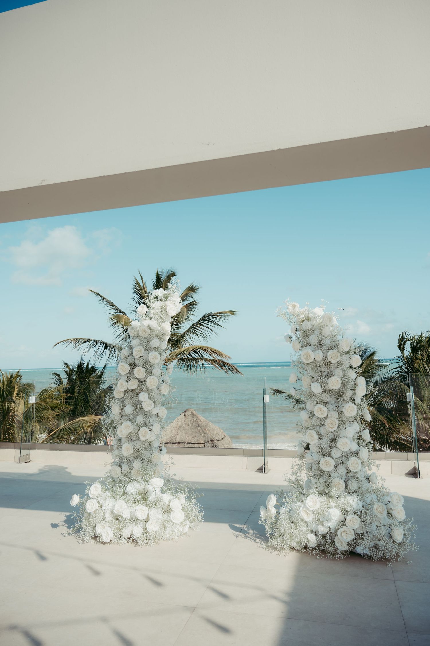 Two elegant white floral arch columns frame a stunning oceanfront wedding ceremony venue with palm trees and turquoise water.
