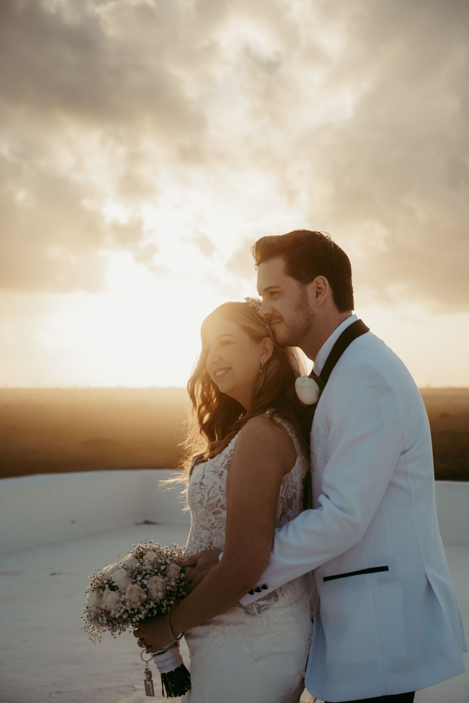 Bride and groom share a romantic sunset kiss on a rooftop, bride holding a white bouquet in wedding attire.