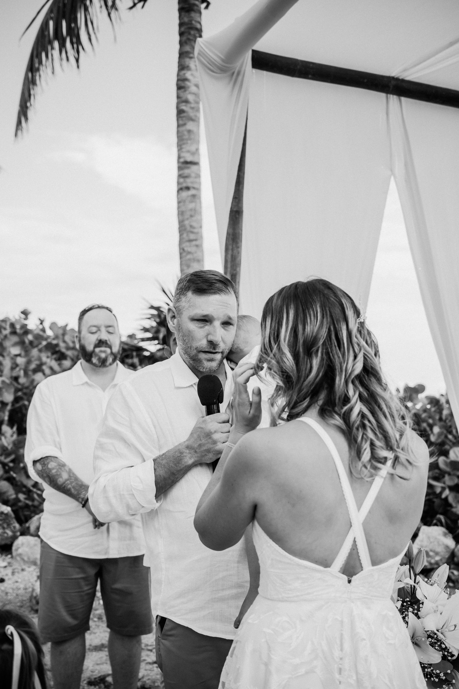 Black and white wedding ceremony photo showing couple exchanging vows under a palm tree with officiant present.
